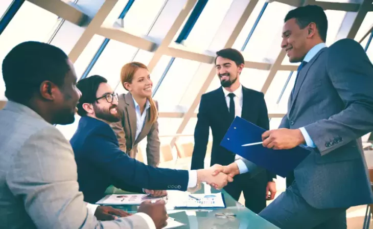 A group of diverse professionals in a modern office space, shaking hands and collaborating over documents, with large windows letting in ample sunlight.